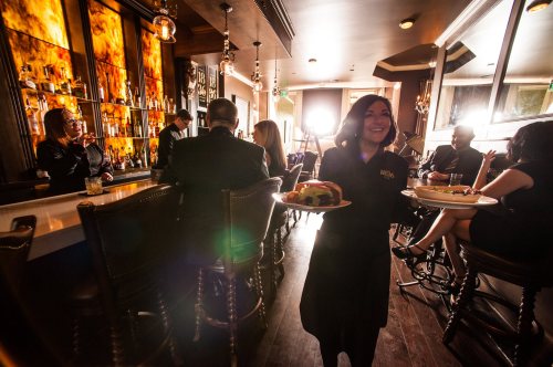 A waitress smiles while serving food in a stylish restaurant with a bar and patrons enjoying their meals.