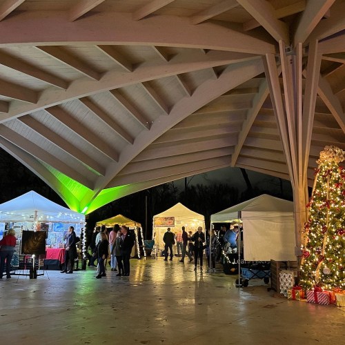 A festive outdoor market under a large pavilion, featuring tents, people, and a decorated Christmas tree.