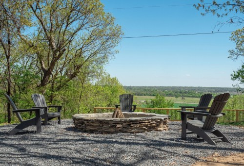 A stone fire pit surrounded by black Adirondack chairs, set against a scenic view of green fields and blue sky.