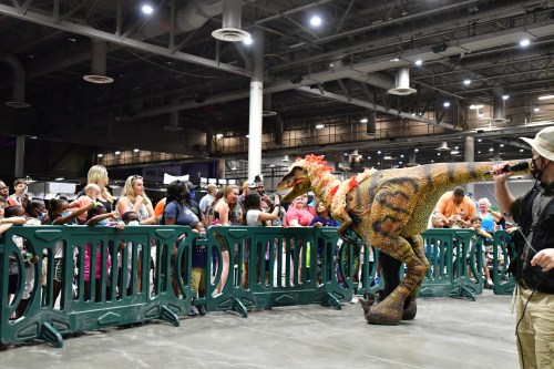 A crowd watches as a person leads a dinosaur costume, adorned with flowers, at an indoor event.