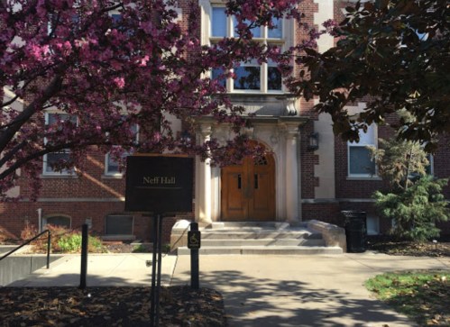 Neff Hall entrance with wooden doors, surrounded by blooming pink trees and greenery on a sunny day.