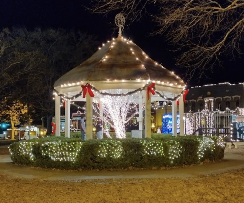 A festive gazebo adorned with lights and ribbons, surrounded by greenery, in a nighttime winter setting.
