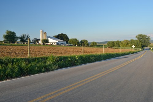 A rural road leads to a farm with silos, fields, and trees under a clear blue sky.
