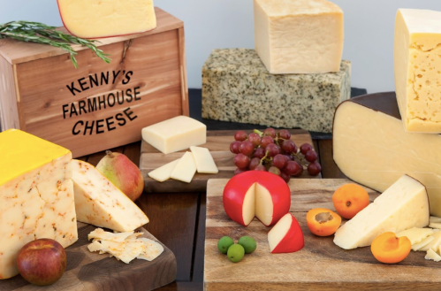 A variety of cheeses displayed on wooden boards, with fruits and a wooden box labeled "Kenny's Farmhouse Cheese."
