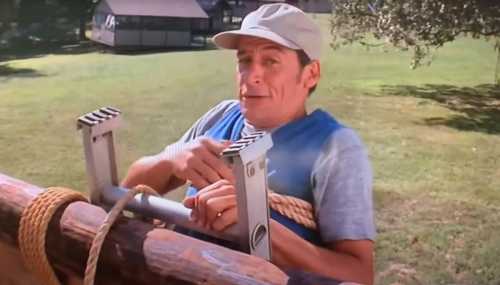 A man in a cap is humorously stuck on a wooden structure, looking surprised while holding onto a rope.