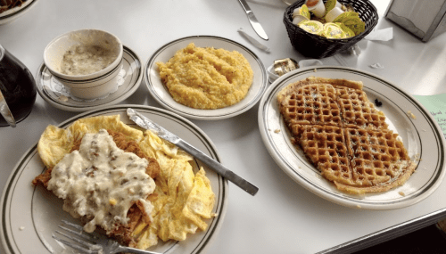 A plate with fried chicken and gravy, scrambled eggs, grits, and a waffle on a table with condiments.