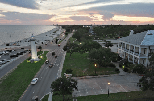 A coastal view at sunset featuring a lighthouse, road, and parked cars along the shoreline.