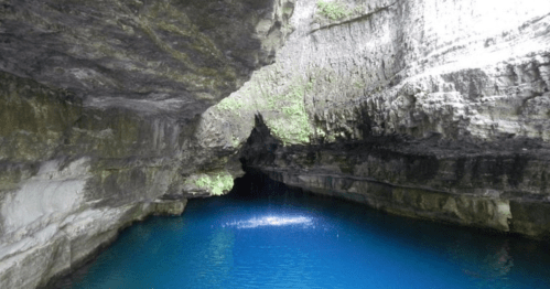 A serene cave with steep rocky walls and a vibrant blue pool of water at the center, illuminated by soft light.
