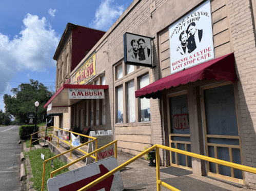 Exterior of a building featuring "Bonnie & Clyde Last Stop Café" and "A.M. Bush" signs, with yellow barriers in front.