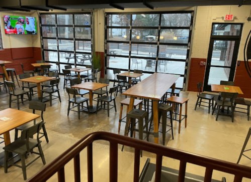 A spacious restaurant interior with wooden tables and chairs, large windows, and a TV displaying a sports game.