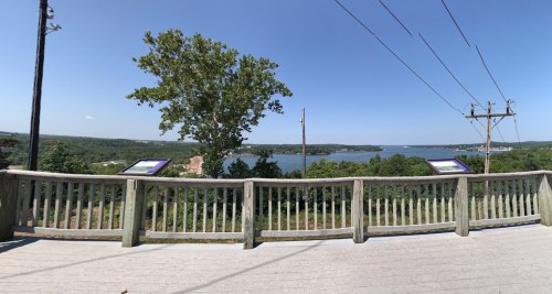 Panoramic view of a lake surrounded by greenery, with informational signs and power lines in the foreground.