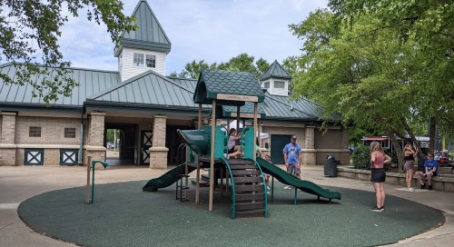 A playground with a green slide and climbing structure, surrounded by trees and a building in the background.