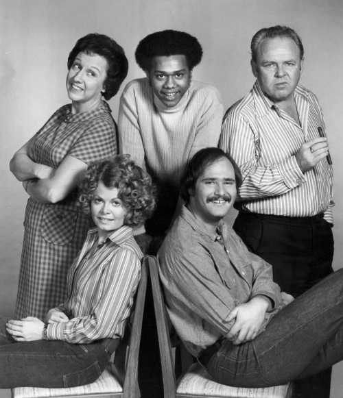 A black and white photo of five actors posing together, smiling and looking at the camera in a casual setting.