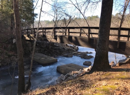 A wooden bridge spans a frozen river, surrounded by trees and rocky banks in a serene winter landscape.