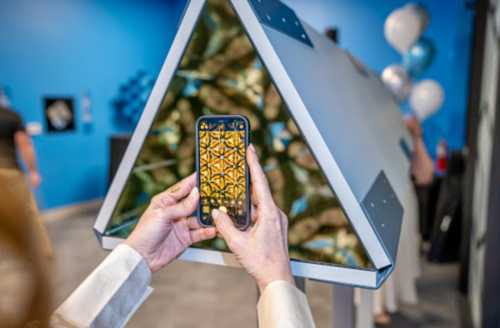 A person holds a phone to capture an image of a triangular display with a colorful pattern, set against a blue wall.