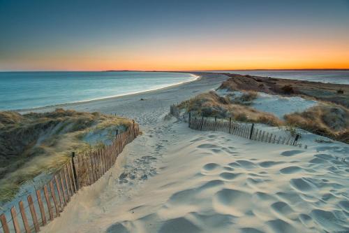 A serene beach scene at sunset, with sand dunes and a wooden fence leading to the calm ocean.