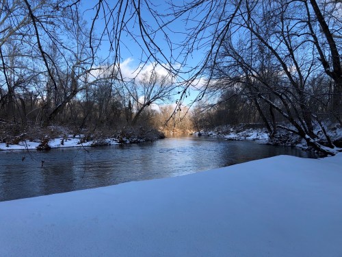A serene winter scene of a river surrounded by snow-covered banks and bare trees under a blue sky.