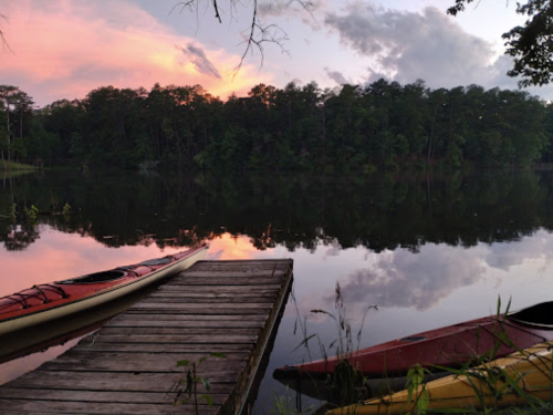 A serene lake at sunset, with kayaks on a wooden dock and trees reflecting in the calm water.