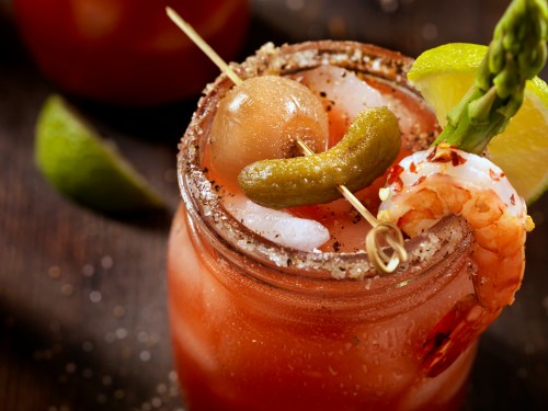A close-up of a cocktail in a jar, garnished with shrimp, olives, pickles, and lime, on a wooden surface.