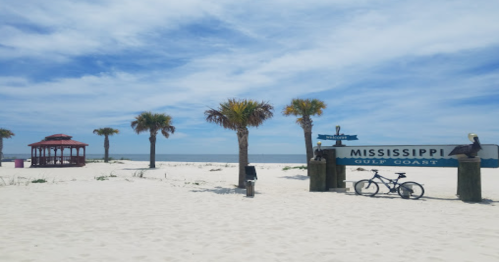 A sunny beach scene with palm trees, a gazebo, and a sign for Mississippi Gulf Coast, with a bicycle nearby.