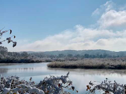 A serene winter landscape featuring a snowy riverbank, calm water, and distant hills under a clear blue sky.