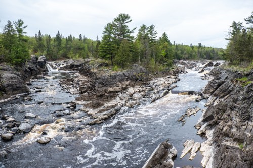 A scenic river flows through rocky terrain, surrounded by lush green trees under a cloudy sky.