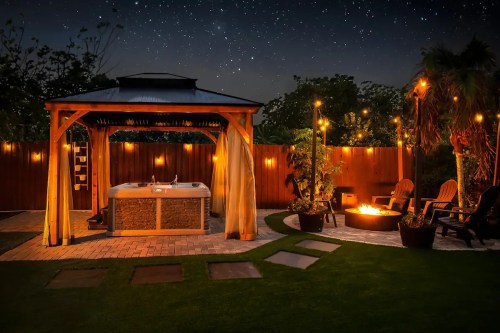 A cozy outdoor space featuring a hot tub, fire pit, and string lights under a starry night sky.