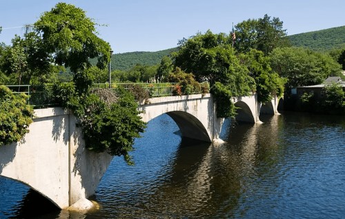A stone bridge with arches spans a calm river, surrounded by lush greenery and hills under a clear blue sky.