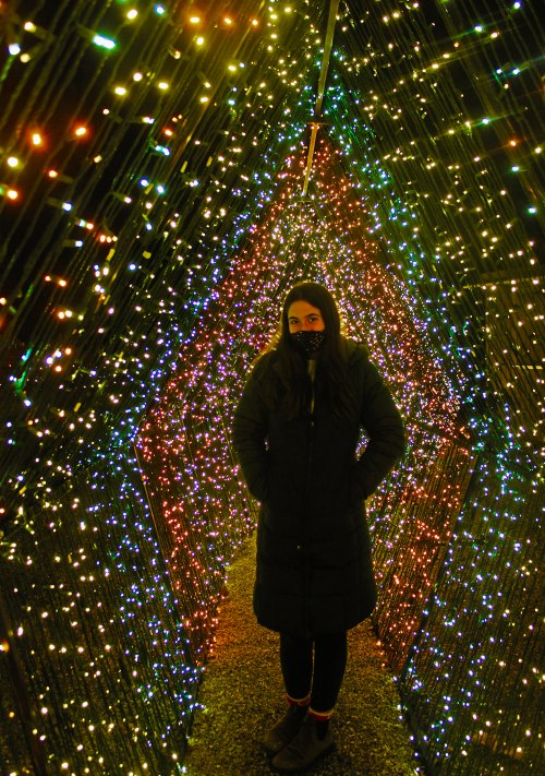 A person stands in a colorful tunnel of lights, wearing a mask and a long coat, surrounded by vibrant, glowing decorations.