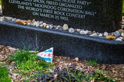 A small Chicago flag rests on the ground near a memorial stone surrounded by rocks and greenery.