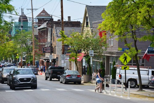 A busy street scene with cars, pedestrians, and shops lined with trees on a sunny day.