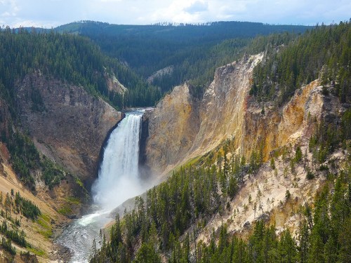 A stunning waterfall cascades down a rocky cliff, surrounded by lush green forests and mountainous terrain.
