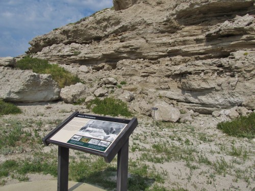 A sign stands in front of a rocky cliff, surrounded by grass and shrubs, under a partly cloudy sky.