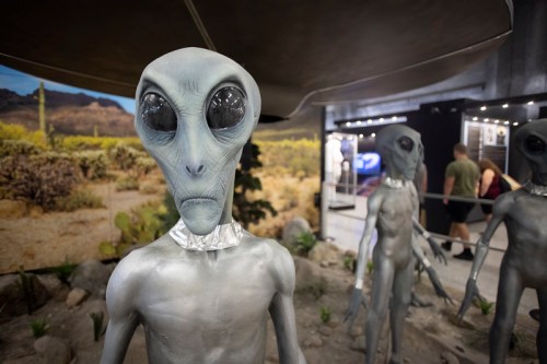 A group of gray alien figures stands under a spaceship, set against a desert landscape backdrop.