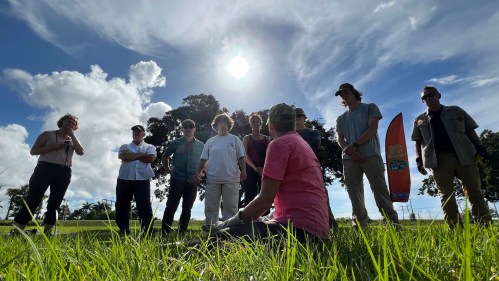 A group of people gathered outdoors, standing and sitting in a circle on grass, with a bright sun overhead.