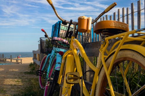 Two colorful bicycles parked by a wooden fence near a sandy beach, with a clear sky and ocean in the background.