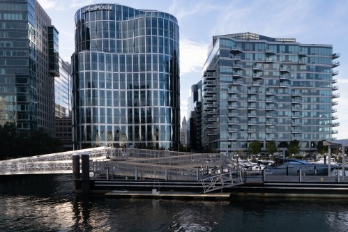 Modern buildings reflect in the water, with a dock and boats in the foreground under a clear blue sky.