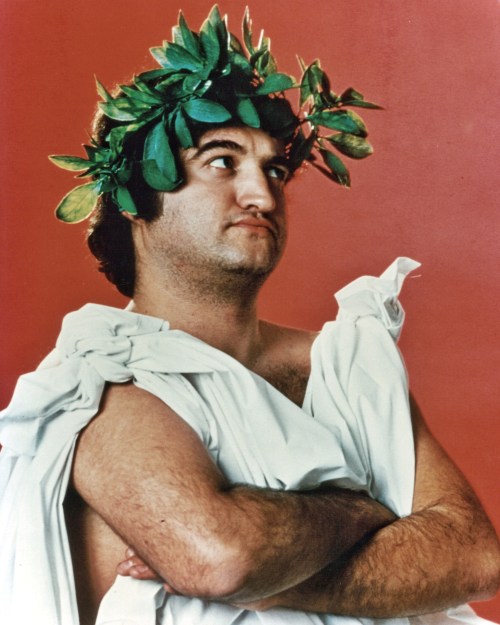 A man in a toga with a laurel wreath poses confidently against a red background.