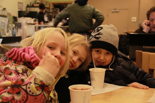 Three children smiling and posing together at a café, with cups in front of them and a cozy atmosphere.