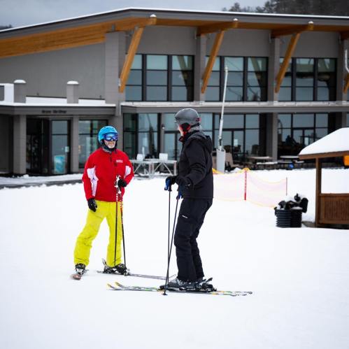 Two skiers in colorful gear stand on snow, chatting near a ski lodge with large windows in the background.