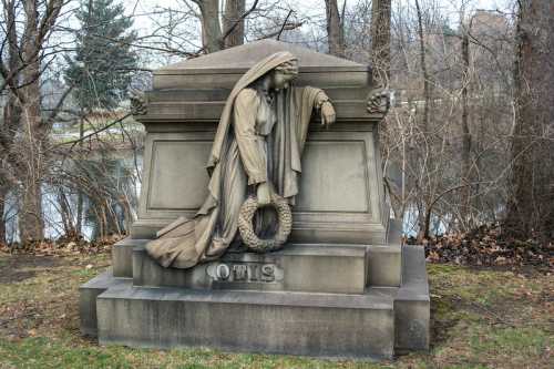 A stone monument featuring a draped figure holding a wreath, surrounded by bare trees and a serene water backdrop.