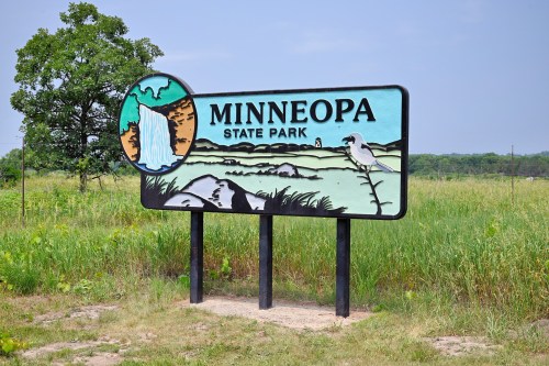 Sign for Minneopa State Park featuring a waterfall illustration and surrounded by green grass and trees.