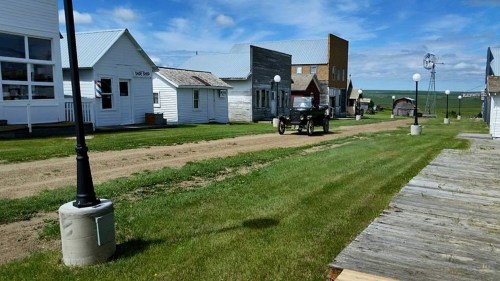 A historic street scene with vintage buildings, a dirt road, and an old car under a blue sky with scattered clouds.
