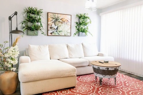 Cozy living room with a white sectional sofa, decorative plants, a wooden coffee table, and a patterned rug.