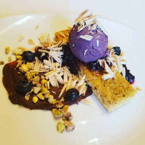 A dessert plate featuring a purple ice cream scoop, chocolate sauce, berries, and almond flakes on a cookie base.