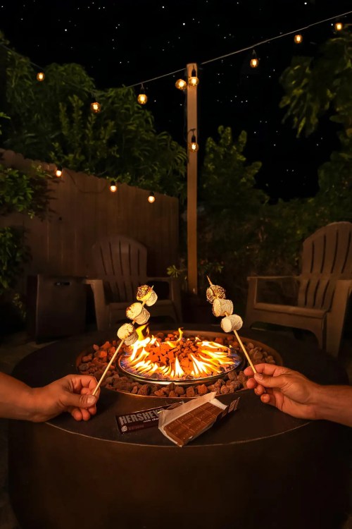 Two hands hold skewers with marshmallows over a glowing fire pit, surrounded by string lights and a starry night sky.