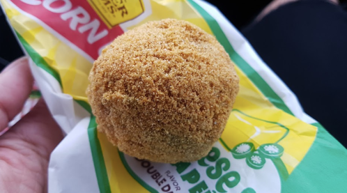 A hand holding a golden, round corn snack coated in a crunchy, textured layer, resting on a colorful packaging.