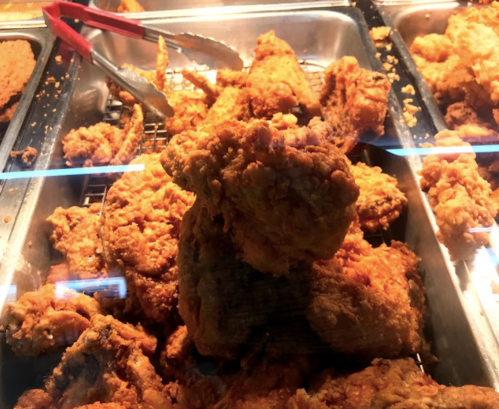 A close-up of crispy fried chicken pieces in a display case, with tongs resting on the side.