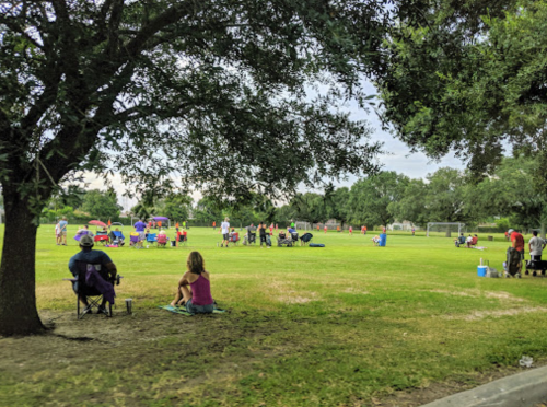 A grassy field with people sitting on chairs, enjoying a community event under trees. Soccer goals in the background.
