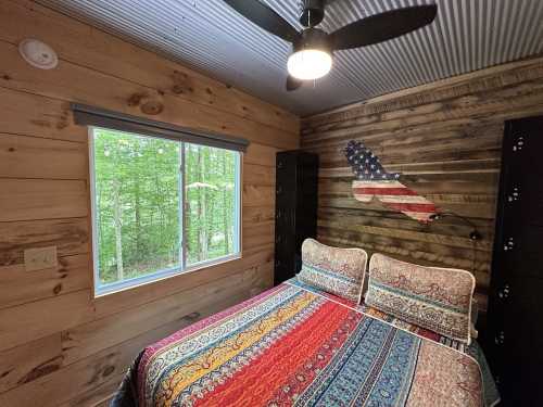 Cozy bedroom with a colorful patterned bedspread, wooden walls, and a window overlooking a green forest.